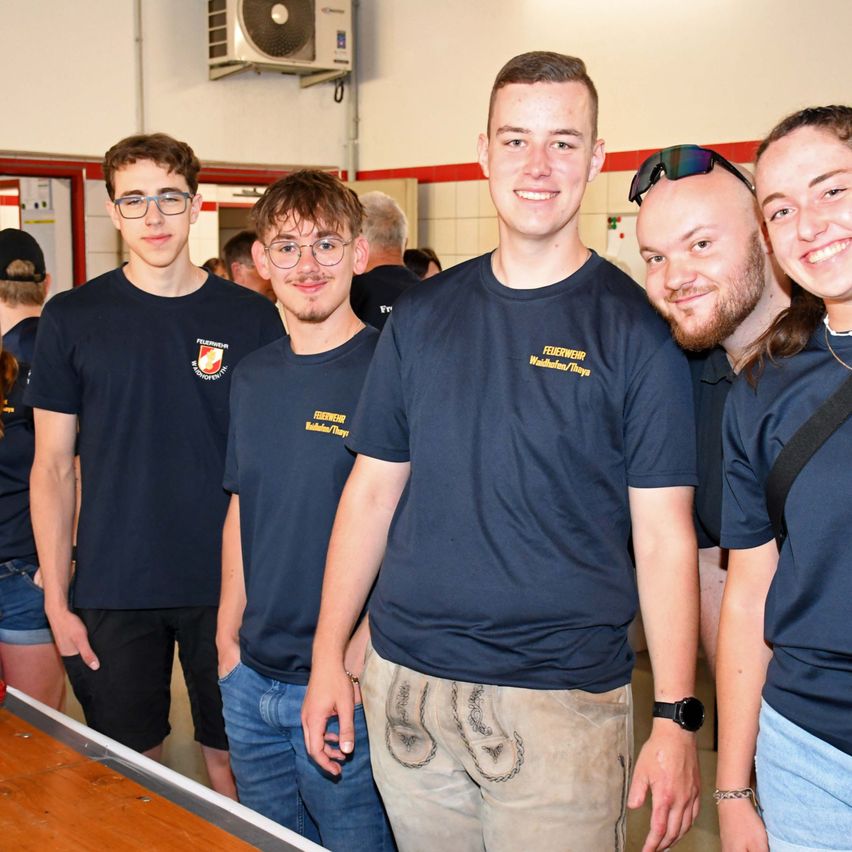 Five young adults in matching navy blue shirts are smiling for a photo. They are standing in a room with a white wall and a red border.