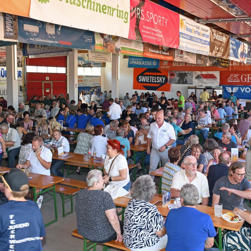 A large group of people are seated at tables in a crowded indoor area, possibly a restaurant or event hall. Many are engaged in conversation, and some are using their phones. The setting is decorated with banners and signs, and the lighting is bright.