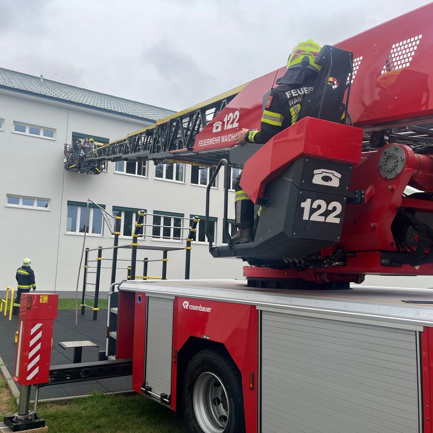 A firefighter is operating a red ladder truck in front of a building. Another firefighter is on the ladder.