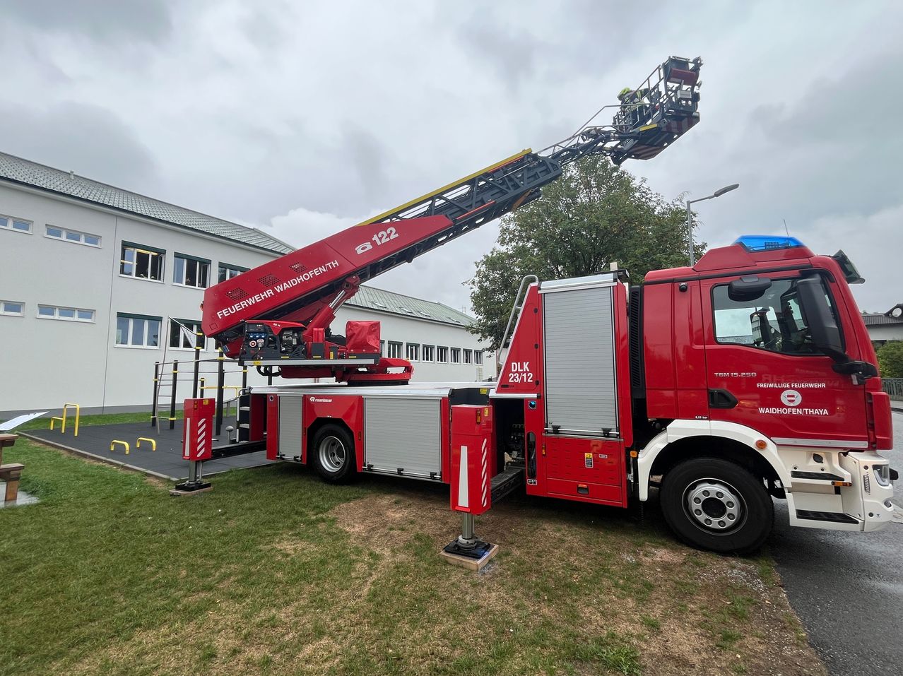 A red fire truck with an extended ladder is parked in front of a building. The ladder is raised and firefighters are on top.