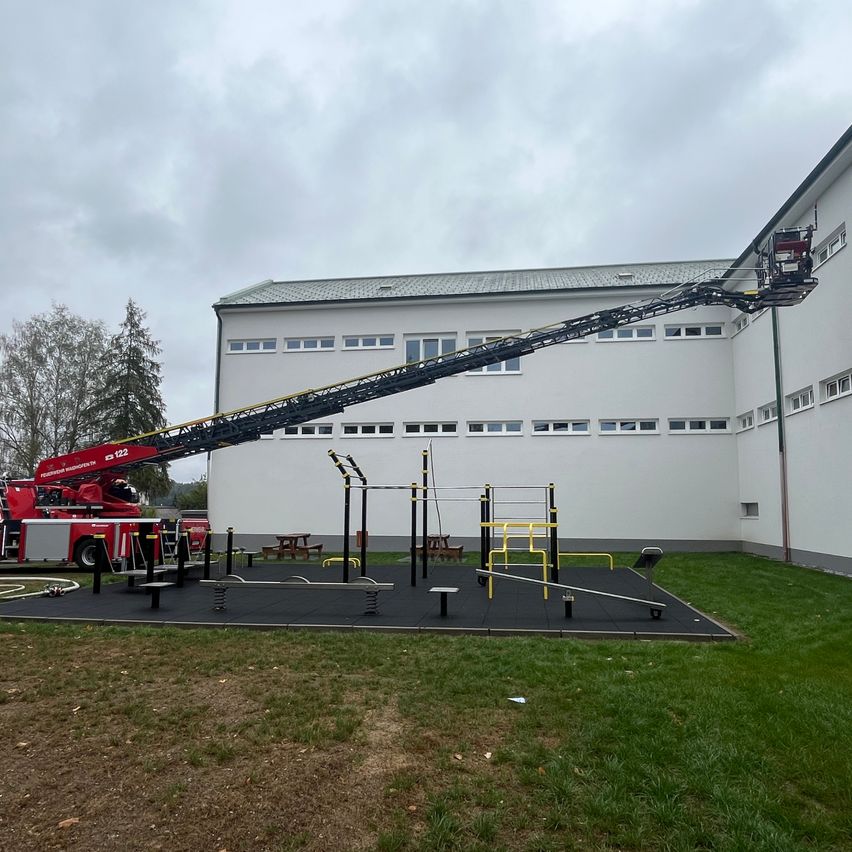 A fire truck with an extended ladder is positioned in front of a white building with several windows. The ladder is reaching towards the building. There are outdoor exercise equipment and benches on the ground.
