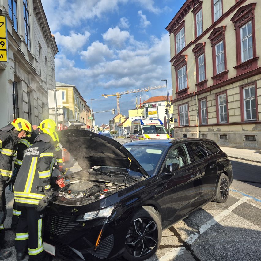Feuerwehrleute inspizieren ein schwarzes Auto mit geöffneter Motorhaube am Straßenrand, das Rauch abgibt. Ein Gebäude mit einem Schild, das 9 liest, ist in der Nähe.