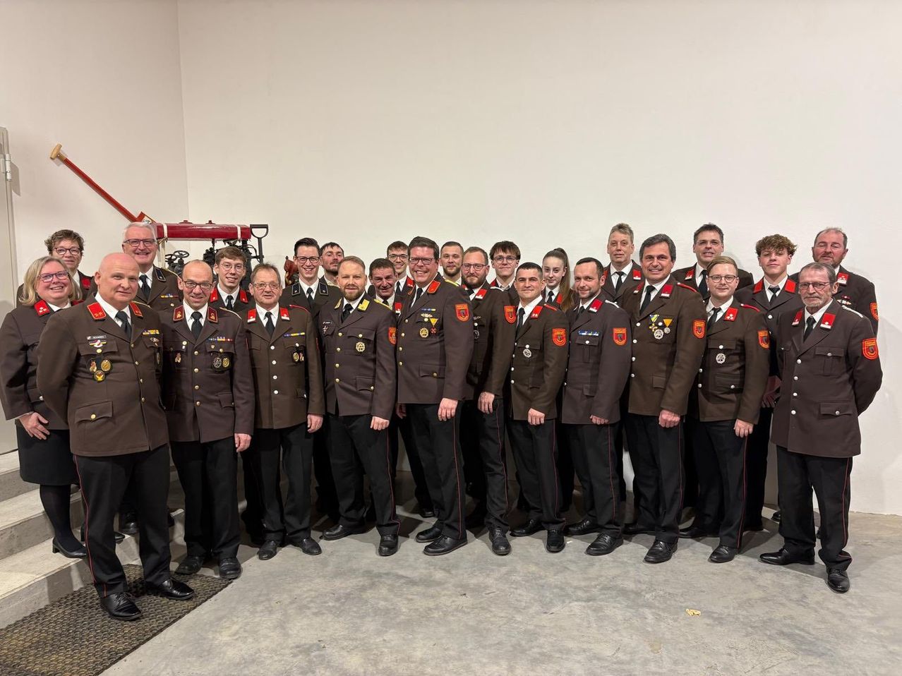 Eine Gruppe uniformierter Personen steht in einer Reihe für ein Foto. Sie tragen braune Uniformen mit roten Streifen und verschiedenen Abzeichen. Der Hintergrund zeigt einen Innenraum mit einer weißen Wand.