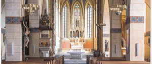 Ein Bild des Kircheninneren mit gewölbten Decken, Buntglasfenstern und Holzbänken. Dekorationen umfassen Statuen und ein Banner mit der Aufschrift 'GEMEINSCHAFT LEBEN' BUNDESWALLFAHRT 2025.