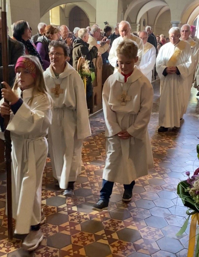 Eine kirchliche Prozession ist im Gange, bei der Kinder und Erwachsene in weißen Gewändern durch den gefliesten Gang einer Kirche gehen. Die Kinder halten einen Stab und eine Blumendekoration.