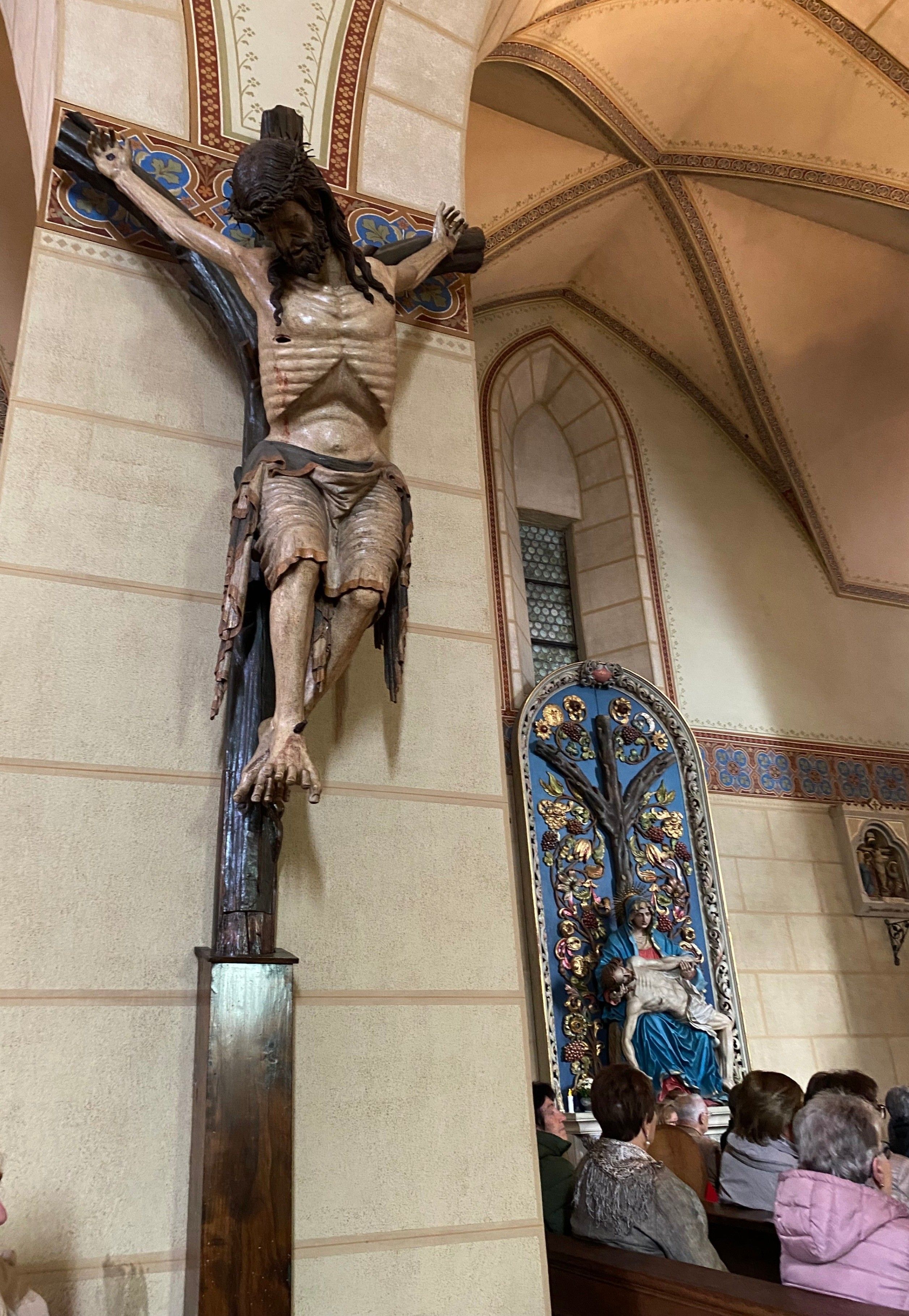 Ein hölzernes Kruzifix mit Jesus am Kreuz an einer Kirchenwand. In der Nähe eine Statue von Maria, die Jesus hält. Unten versammelt sich eine Gruppe von Menschen.