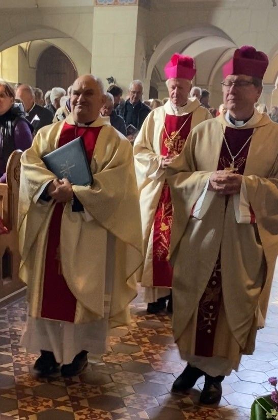 Drei Priester stehen in einer Kirche mit prächtiger Architektur. Einer hält ein Buch, während die anderen ihre Hände zusammengelegt haben. Hinter ihnen stehen mehrere Menschen, darunter eine Frau in einer lila Jacke.