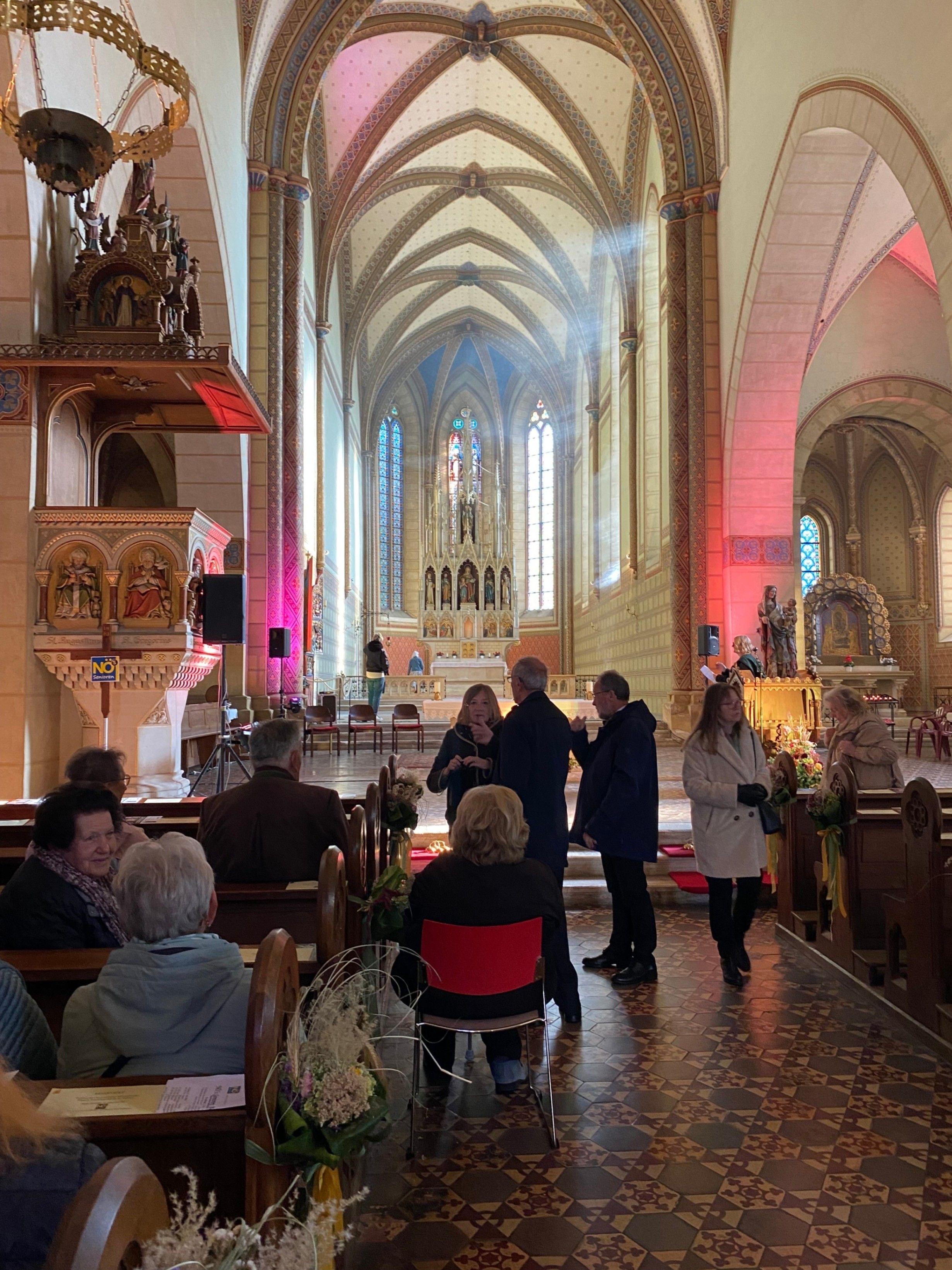 In einer Kirche sind mehrere Menschen versammelt. Einige stehen und unterhalten sich in der Nähe des Altars, während andere auf Bänken sitzen. Dekorationen und Blumen schmücken die Kirchenbänke.