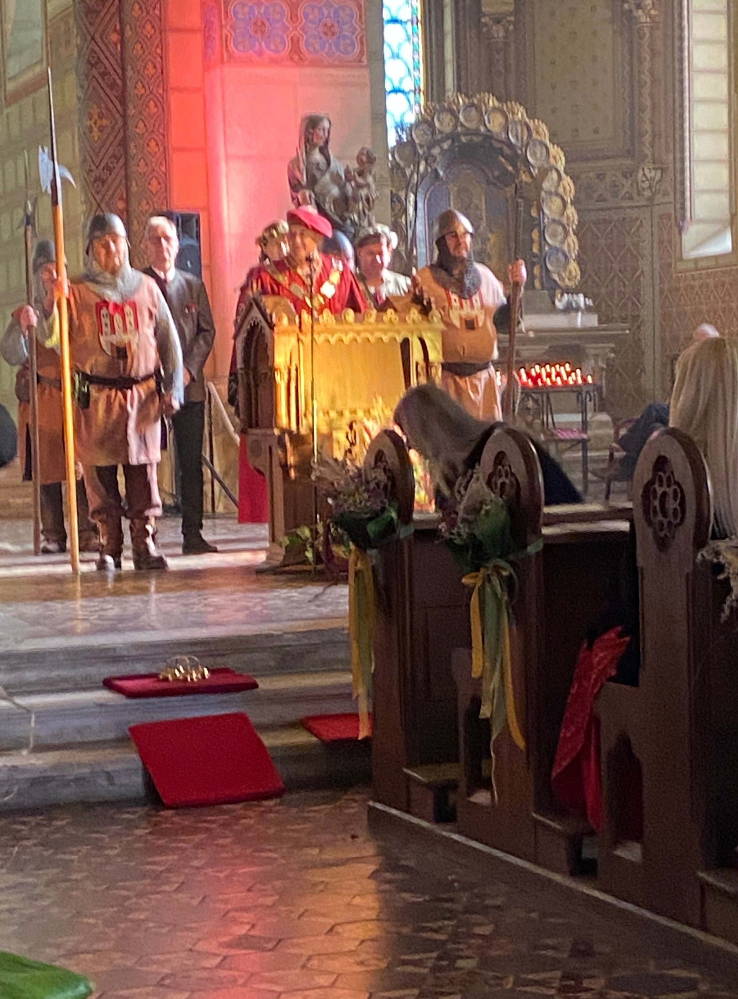 In einer Kirche steht eine Gruppe von Menschen in mittelalterlicher Kleidung neben einem Podium mit Kerzen und Blumendekoration. Einige halten Speere.