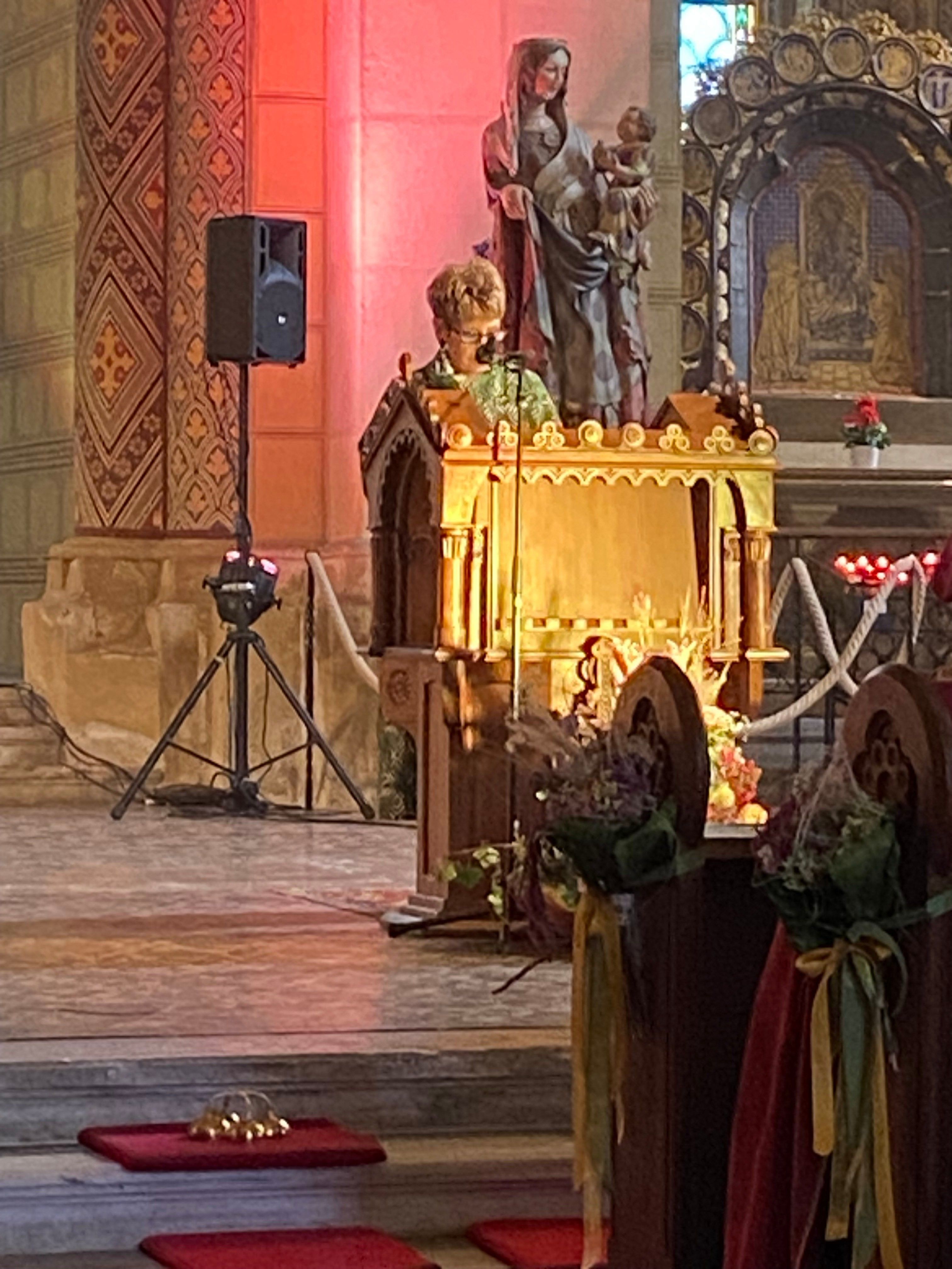 Eine Frau spricht an einer mit Lichtern beleuchteten Kanzel mit einem Lautsprecher in einer Kirche mit einer Statue und einer Kerze hinter ihr.