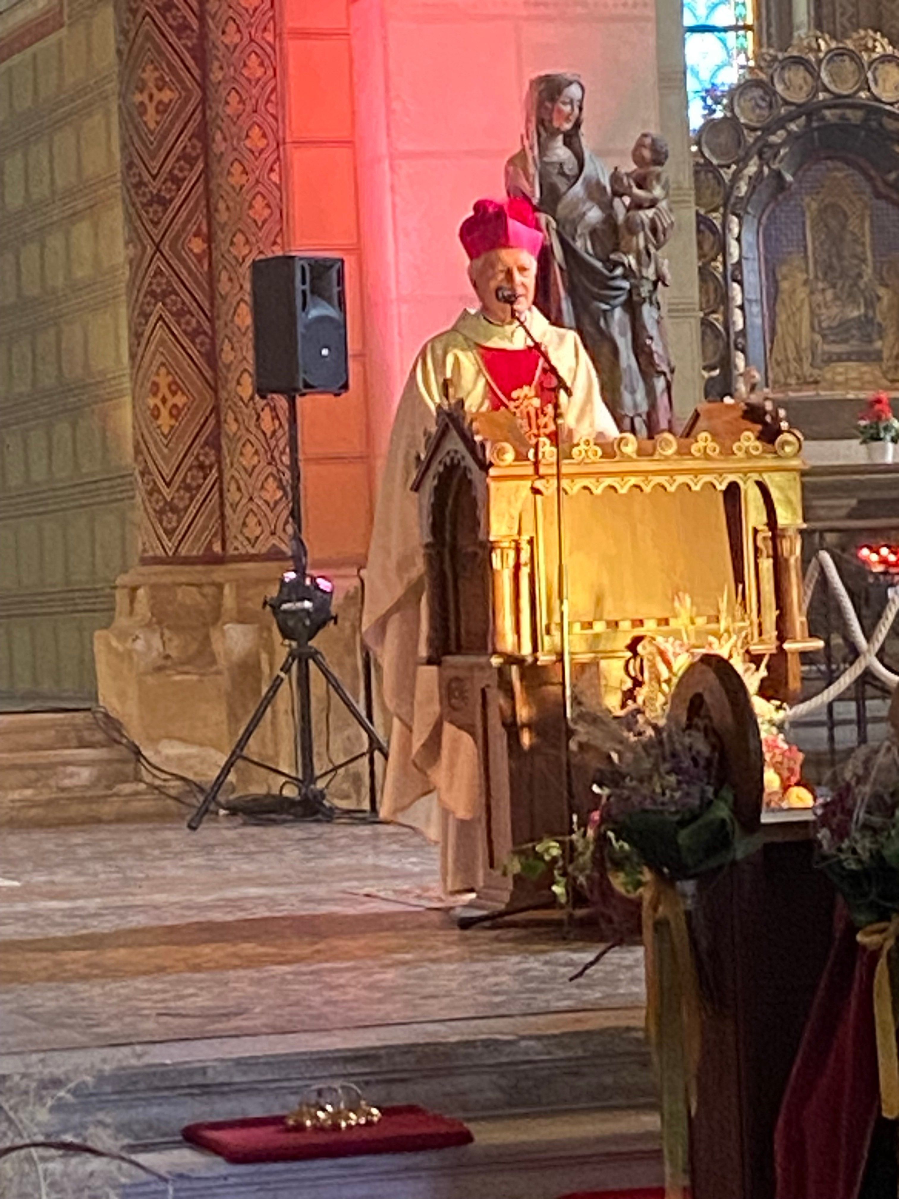 Ein Bischof in einer roten und goldenen Robe steht in einer Kirche an einem Podium, mit einem schwarzen Lautsprecher und Stativ in der Nähe.
