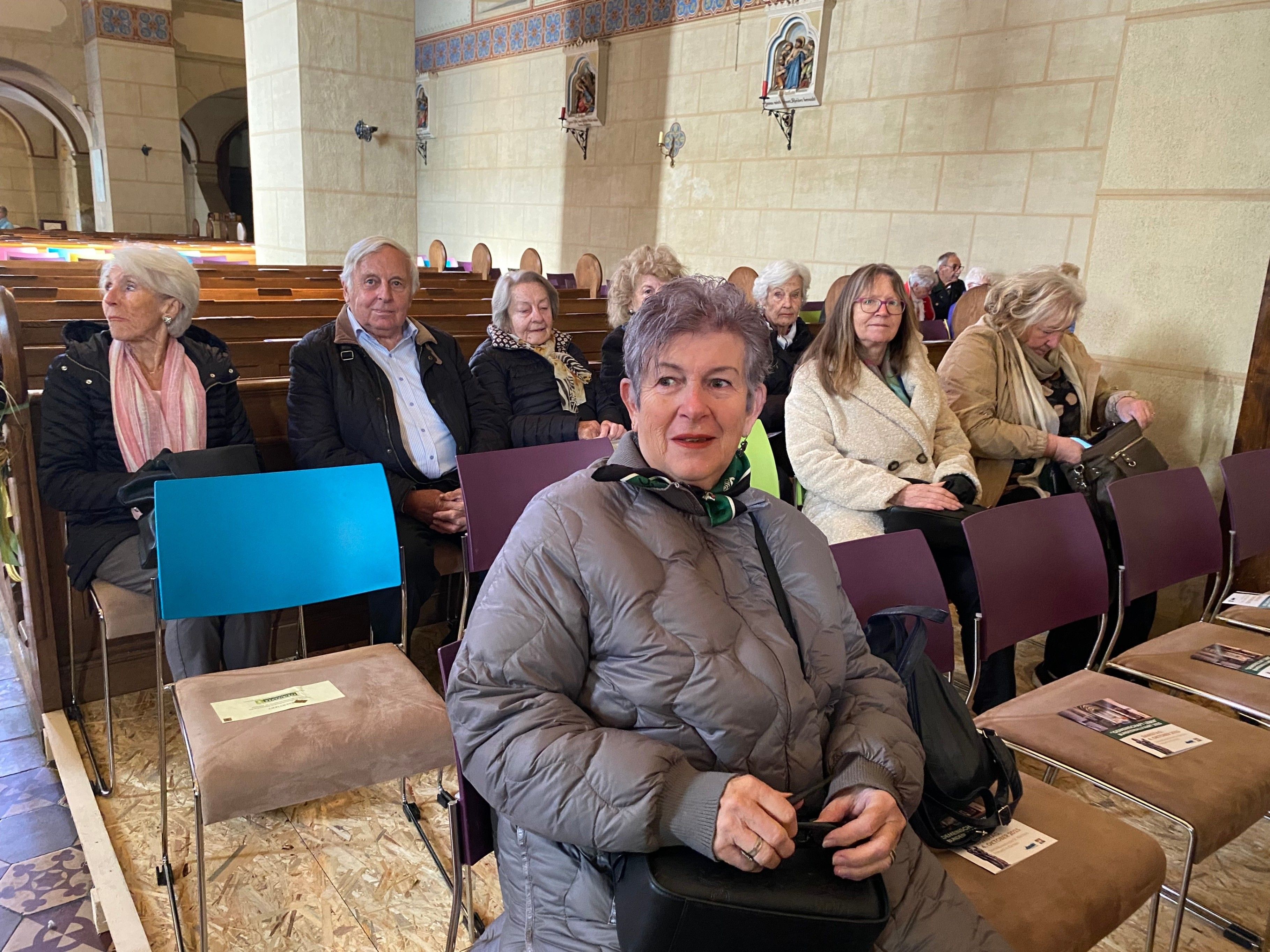 Eine Gruppe älterer Menschen sitzt in einer Kirche. Eine ältere Frau mit kurzen Haaren sitzt im Vordergrund, in einer grauen Jacke. Hinter ihr sind andere warm gekleidet und sitzen in Reihen von Kirchenbänken.