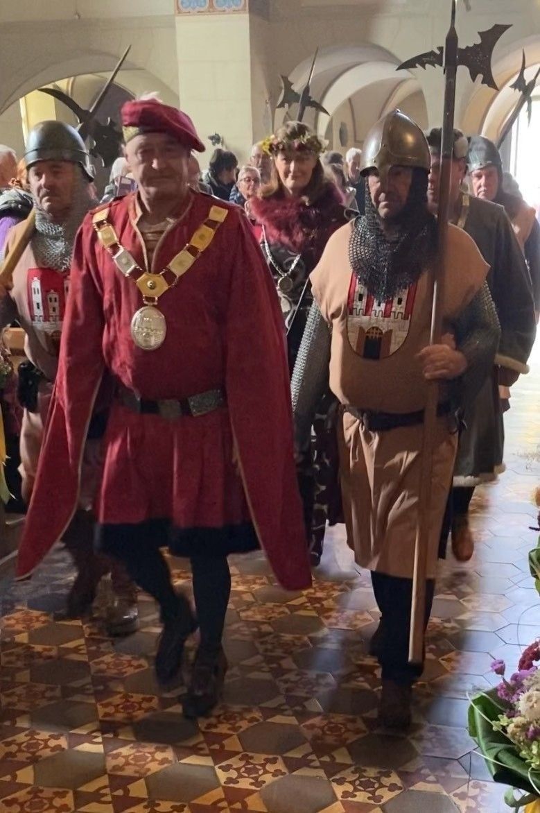 Eine Gruppe von Menschen in mittelalterlichen Kostümen bewegt sich in einer Prozession. Einige tragen Helme, und einer hält eine Lanze. Ein Mann vorne trägt einen roten Umhang und eine goldene Kette.