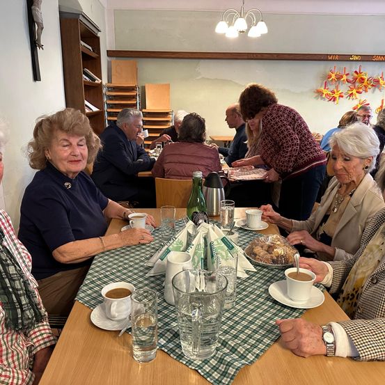 Eine Gruppe älterer Menschen trifft sich in einem Restaurant, um an einem Tisch mit Tellern voller Essen und Tassen Kaffee zu sitzen. Eine Frau steht am Tisch und serviert sie.