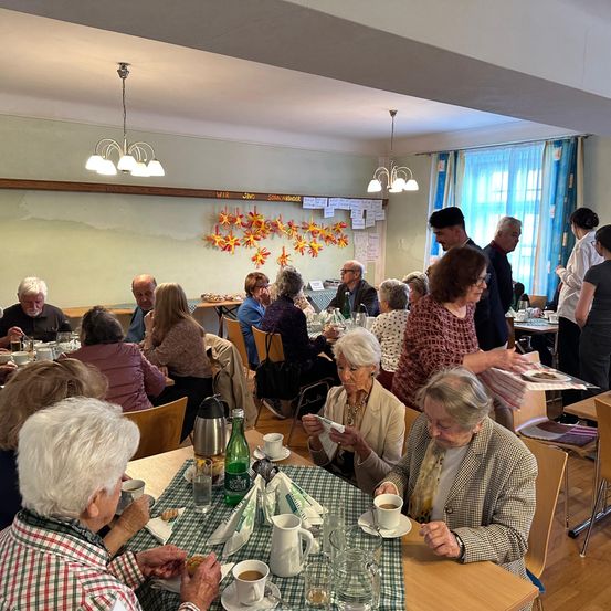 Eine Gruppe älterer Menschen versammelt sich in einem Esszimmer und sitzt an Tischen. Einige lesen oder unterhalten sich, während andere essen. Der Raum ist mit Stühlen, Tischen und dekorativen Gegenständen eingerichtet.
