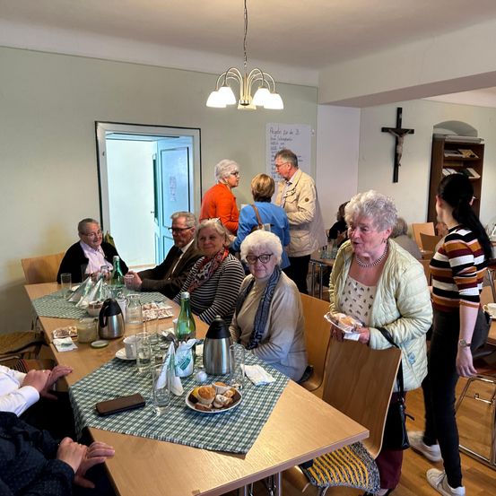 Eine Gruppe älterer Menschen sitzt in einem Esszimmer um einen Tisch herum und isst. Eine Frau steht und hält einen Teller mit Essen. Flaschen, Gläser und andere Gegenstände stehen auf dem Tisch. Eine Frau mit Brille sitzt auf einem Stuhl.