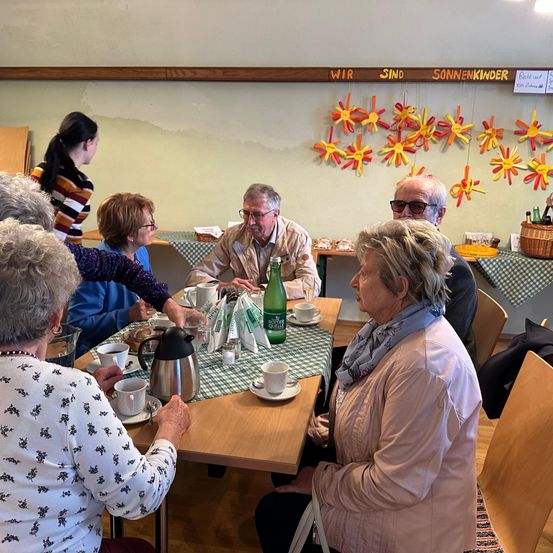 Eine Gruppe älterer Menschen versammelt sich in einem Raum, sitzt um einen Tisch mit Tassen und Tellern herum. Eine Frau in einem Blumenhemd schenkt Kaffee ein. Hinter ihnen steht eine Frau in einem gestreiften Shirt.