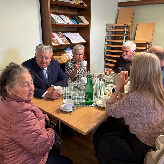 Eine Gruppe von Menschen sitzt um einen Tisch herum, wahrscheinlich in einer Besprechung oder Diskussion. Der Tisch hat Tassen, eine grüne Flasche und ein grünes Glas. Dahinter steht ein Bücherregal aus Holz mit Büchern und Papieren.
