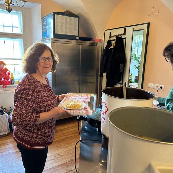 Eine Frau in der Küche, die einen Teller mit Essen in der Hand hält, steht neben großen Töpfen. Eine andere Person ist im Hintergrund.