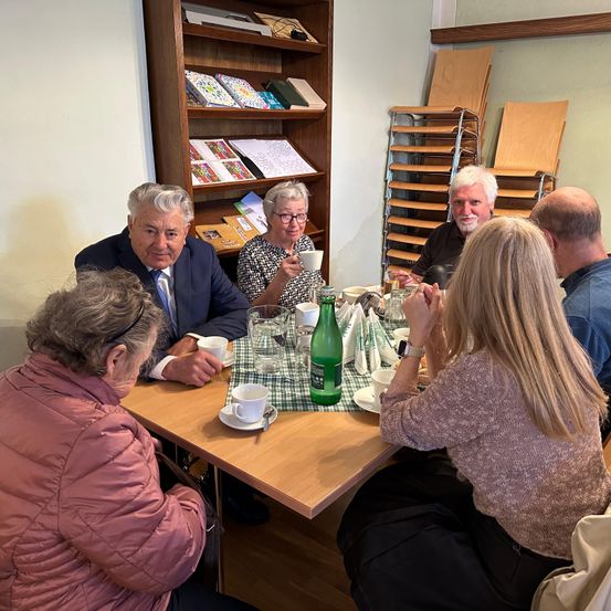 Eine Gruppe von Menschen sitzt um einen Tisch in einem Raum und isst und trinkt. Ein Mann in Anzug schaut eine Frau mit Brille an.