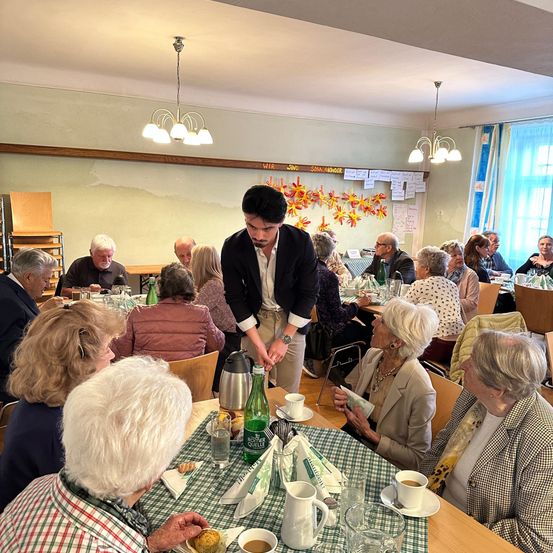Ein Mann serviert Kaffee an einem Tisch in einem Esszimmer. Ältere Menschen sitzen um die Tische herum, essen und trinken. Auf dem Tisch stehen eine grüne Flasche und eine Karaffe.