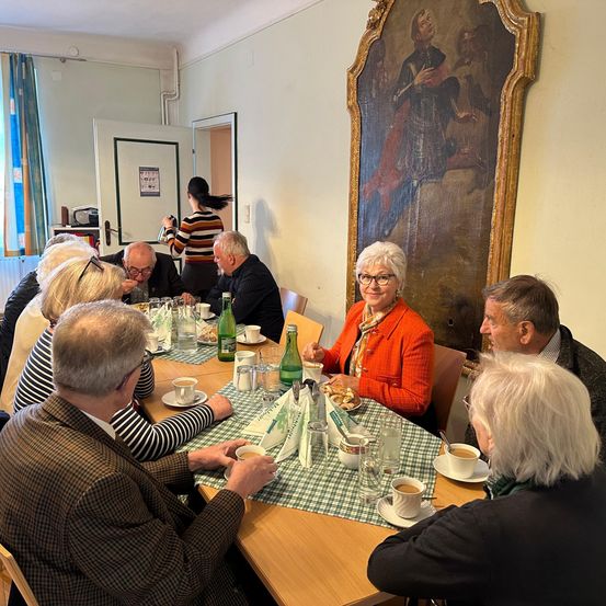 Eine Gruppe von Menschen sitzt um einen Tisch herum und führt ein Gespräch. Eine Frau in einer orangefarbenen Blazerjacke lächelt und scheint mit einer anderen Person zu sprechen. Der Tisch ist mit Tassen, Tellern und Servietten gedeckt. Ein Gemälde hängt an der Wand hinter ihnen.
