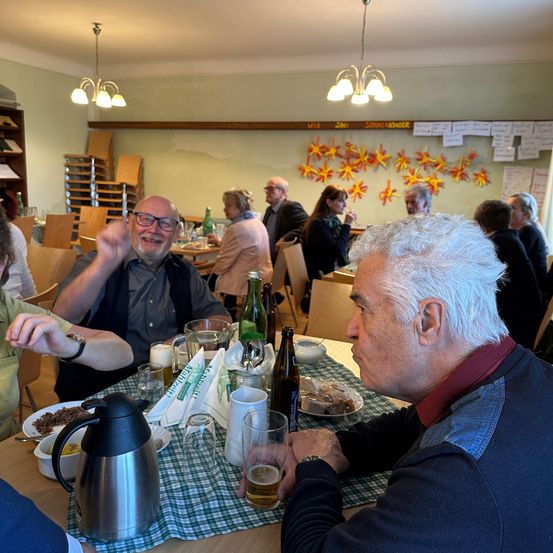 Mehrere Personen sitzen in einem Restaurant an einem Tisch und unterhalten sich. Der Tisch ist mit Tellern, Gläsern und einer Karaffe Bier gedeckt. Ein Mann hat eine Tasse Kaffee, und ein anderer Mann lächelt in die Kamera.