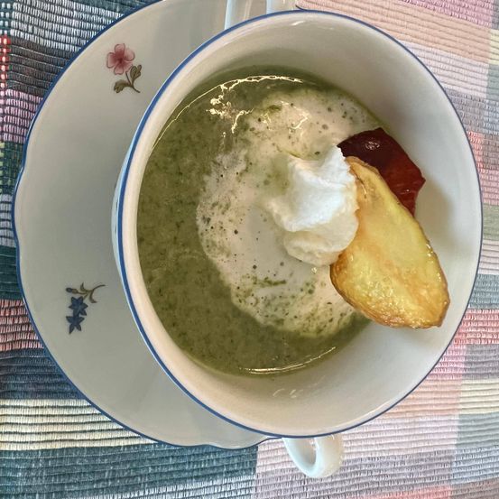 Eine Schüssel grüner Suppe mit einem Stück Zitrone und Schlagsahne oben drauf. Die Suppe steht auf einer Untertasse mit einem Blumendesign.