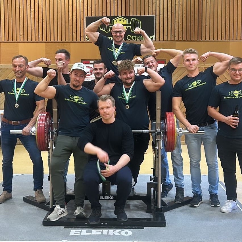 Eine Gruppe von Männern in passenden T-Shirts posiert mit Hanteln in einem Fitnessstudio für ein Foto. Sie haben Medaillen um den Hals.