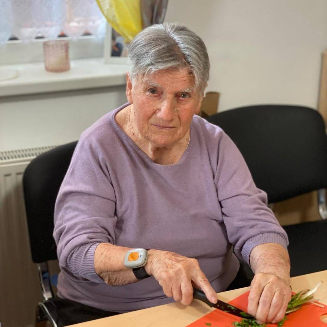 Eine ältere Frau in einem lila Langarmshirt schneidet Gemüse auf einem Tisch.