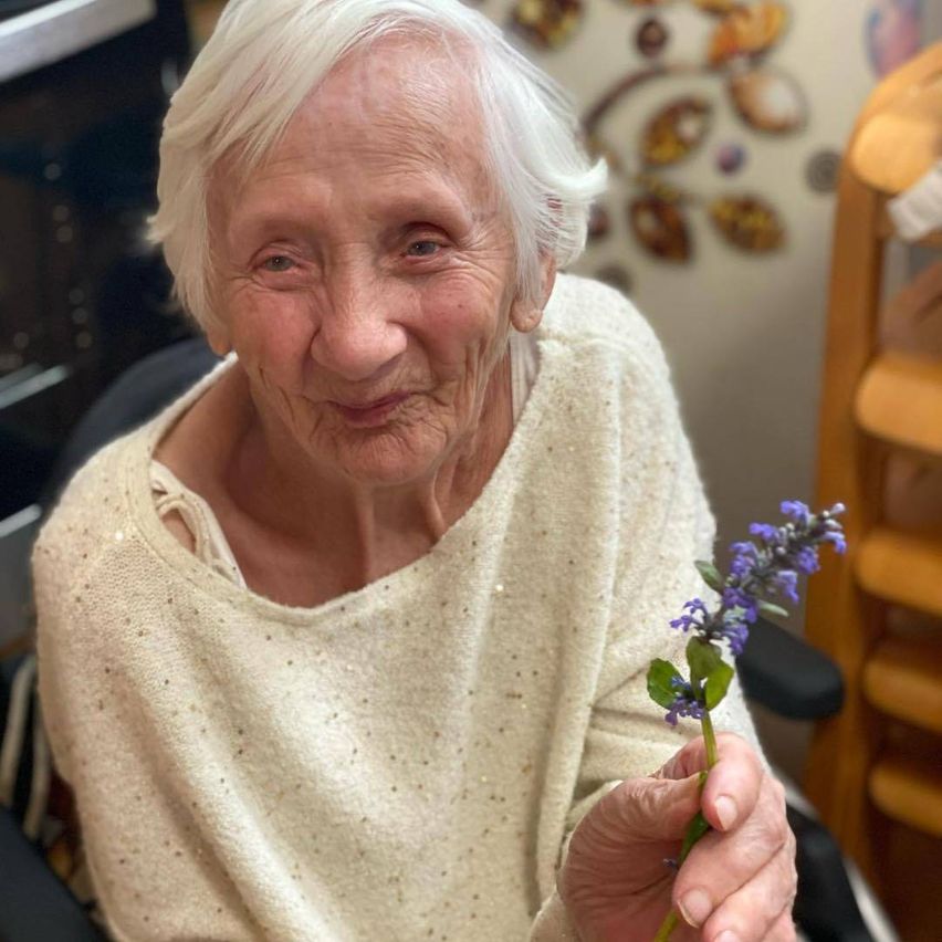 Eine ältere Frau mit weißen Haaren sitzt in einem Stuhl. Sie lächelt und hält eine Blume in der Hand. Sie trägt einen beigefarbenen Pullover.