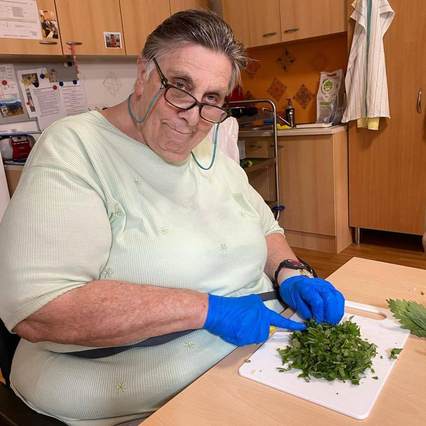 Eine Frau in einem hellgrünen Shirt, mit Brille und blauen Handschuhen schneidet Kräuter auf einem weißen Schneidebrett in einer Küche.