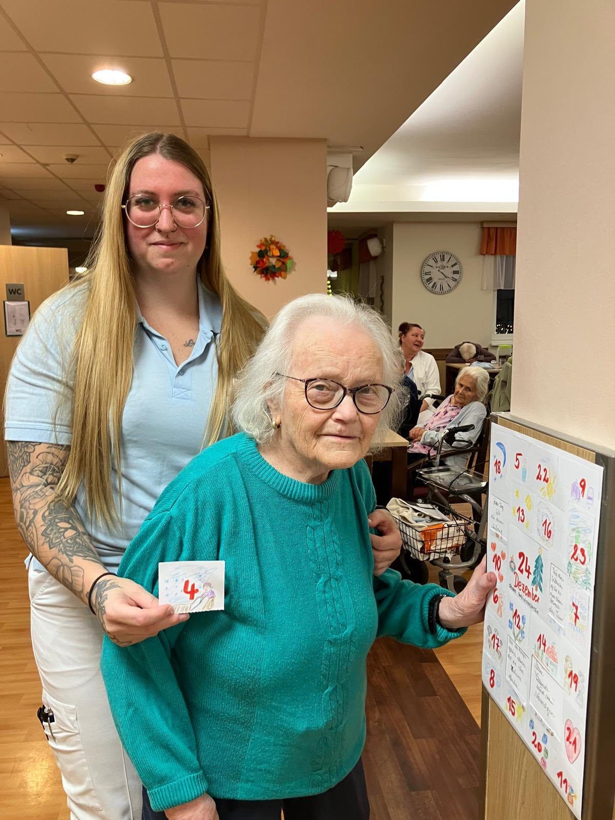 Zwei Frauen stehen in einem Raum mit einem Kalender. Die Frau auf der linken Seite hält ein Papier mit der Nummer vier. Die Frau auf der rechten Seite schaut auf den Kalender.