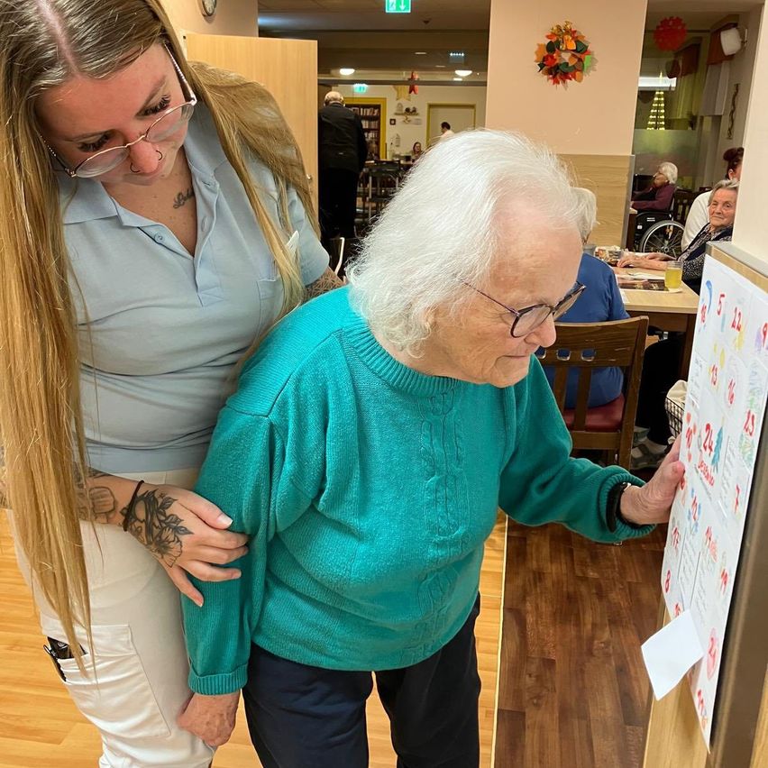 Eine ältere Frau mit weißen Haaren, die einen türkisfarbenen Pullover trägt, schaut auf einen Kalender. Sie wird von einer Frau mit Brille und Tätowierungen am Arm unterstützt.