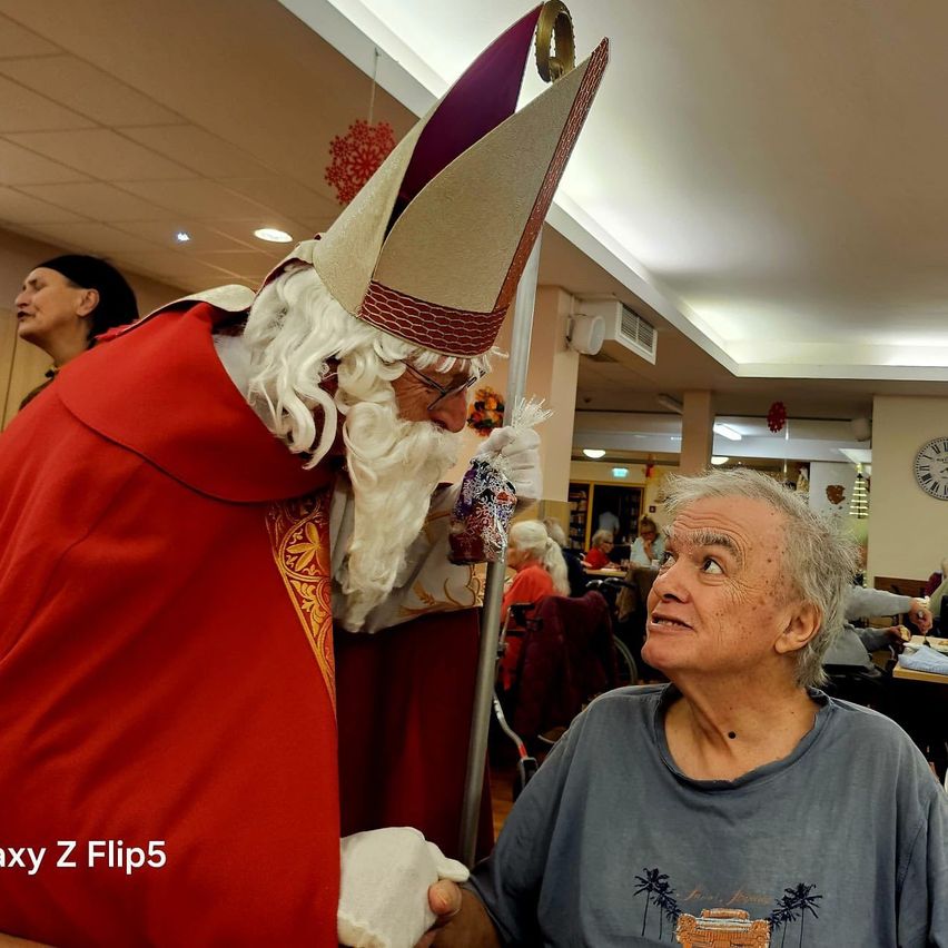 Ein Mann, der als Weihnachtsmann verkleidet ist, steht neben einer älteren Frau in einem Restaurant. Der ältere Mann sieht überrascht aus. Hinter ihnen sitzen Menschen an Tischen. Das Bild hat einen Wasserzeichen.