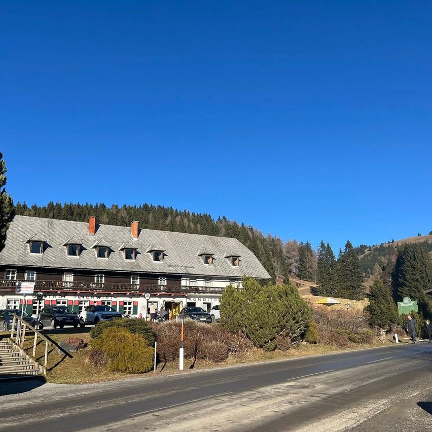 Ein großes Gebäude mit grauem Dach und Schornsteinen steht auf einem Hügel, umgeben von Bäumen. Mehrere Autos sind vor dem Gebäude geparkt. Der Himmel ist klar und blau.