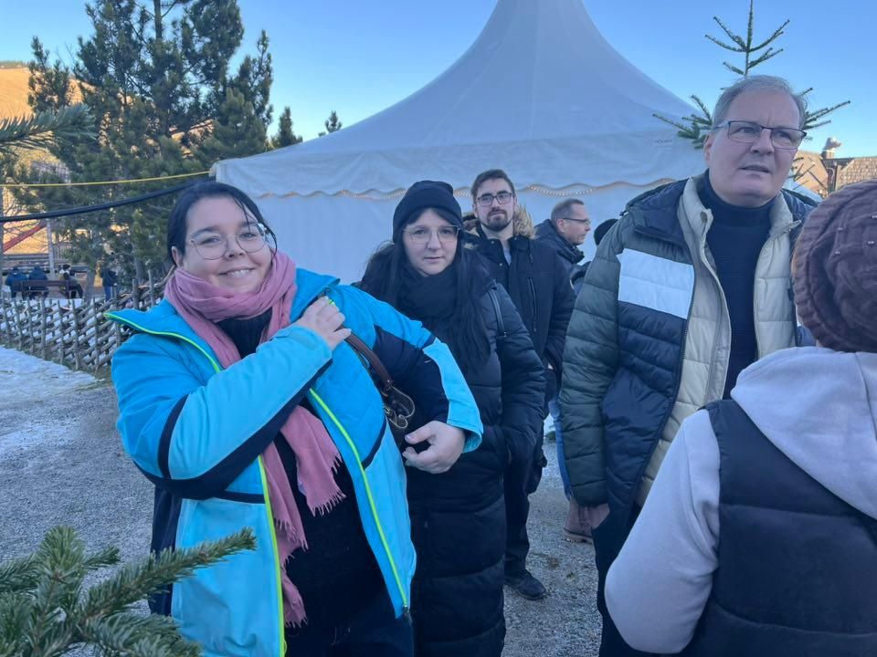 Eine Gruppe von Menschen steht neben einem Weihnachtsbaum in einer winterlichen Umgebung, mit einem großen weißen Zelt im Hintergrund.