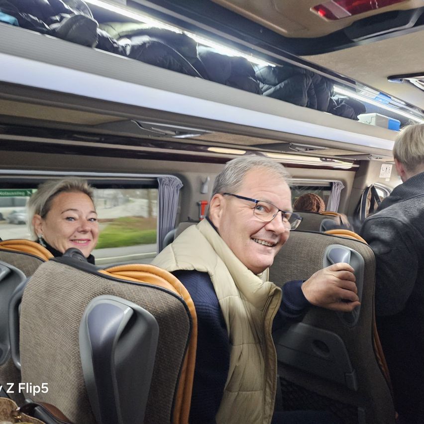 Der Innenraum eines Busses mit zwei Passagieren in der Mitte, eine lächelnde Frau und ein lachender Mann. Über ihnen befinden sich Taschen auf dem Gepäckträger.
