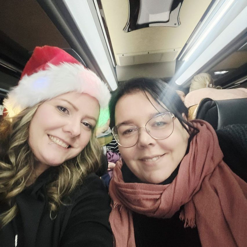 Zwei Frauen in Winterkleidung lächeln für ein Selfie im Bus, eine in einer Weihnachtsmütze und die andere mit Brille und Schal.