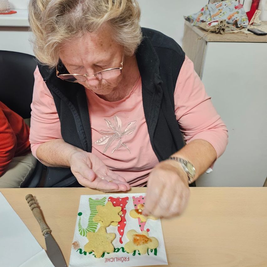 Eine ältere Frau in einem rosa Shirt und einer schwarzen Weste dekoriert Lebkuchen mit Zuckerguss auf einem Holztisch.