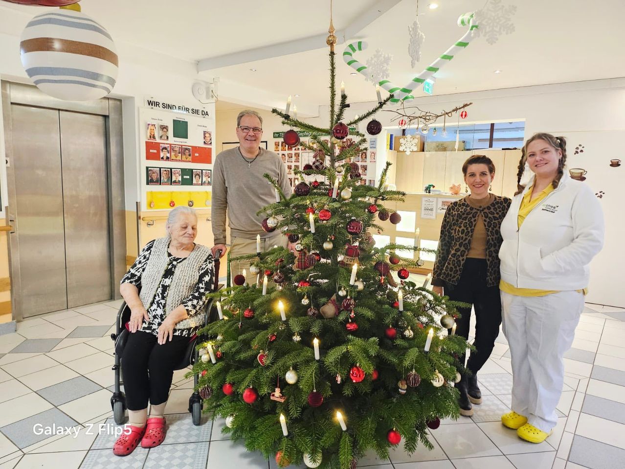 Eine Gruppe von Menschen steht um einen geschmückten Weihnachtsbaum in einem Flur herum. Eine ältere Frau im Rollstuhl lächelt, und ein Mann steht hinter dem Baum.
