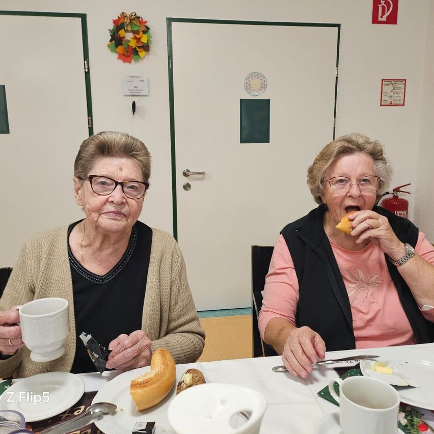 Zwei ältere Frauen sitzen an einem Tisch, eine mit einer Tasse und einem Stück Brot, die andere isst lächelnd. Hinter ihnen befinden sich zwei weiße Türen und eine Girlande an der Wand.