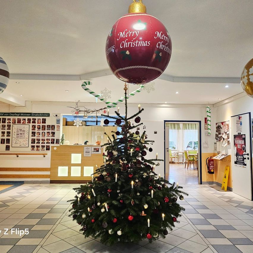 Ein Weihnachtsbaum mit einem großen roten Kugelschmuck oben steht in einer Lobby. Er hat Dekorationen und Kerzen. Dahinter steht ein Schreibtisch mit einem Schild und Fotos. Im Hintergrund sind Stühle und ein Feuerlöscher.