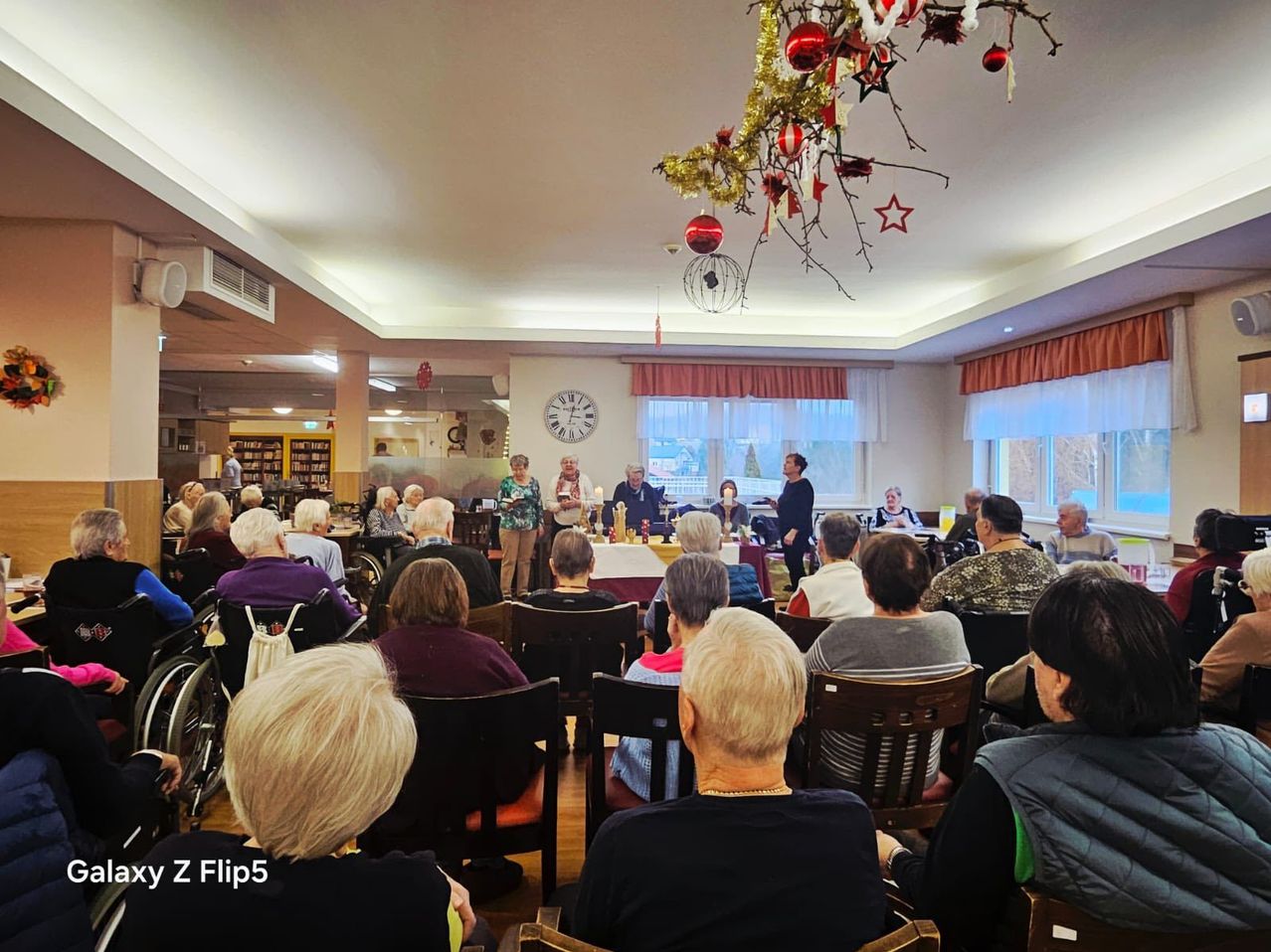 Eine Gruppe älterer Menschen sitzt in einem Raum mit Weihnachtsdekoration, einer Uhr und einem Tisch mit Kerzen in der Mitte. Einige sitzen in Rollstühlen.