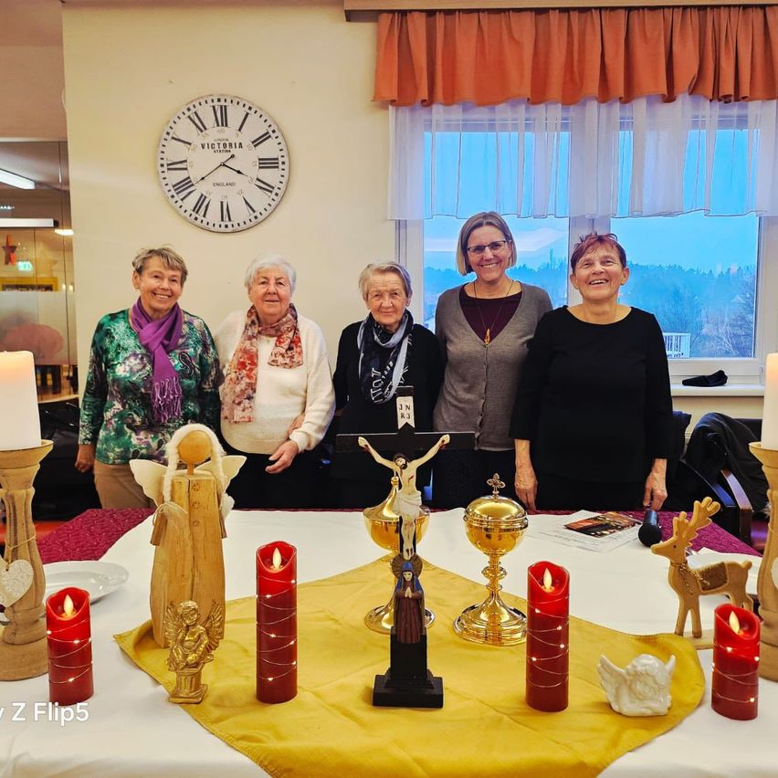 Fünf Frauen stehen lächelnd hinter einem Tisch mit religiösen Gegenständen. Der Tisch hat ein Kreuz, Kerzen und Figuren. Hinter ihnen befindet sich eine Wand mit einer Uhr und einem Fenster.
