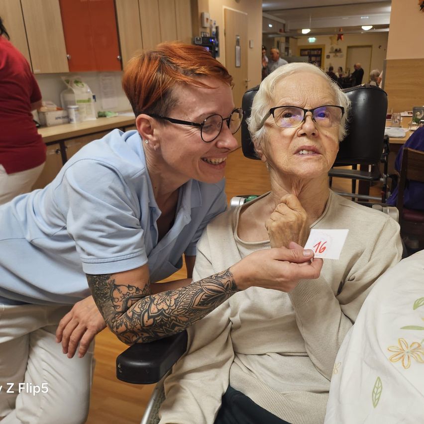 Eine junge Frau hält eine Karte mit der Nummer 16, während sie eine ältere Frau mit Brille und einem beigen Pullover anlächelt.