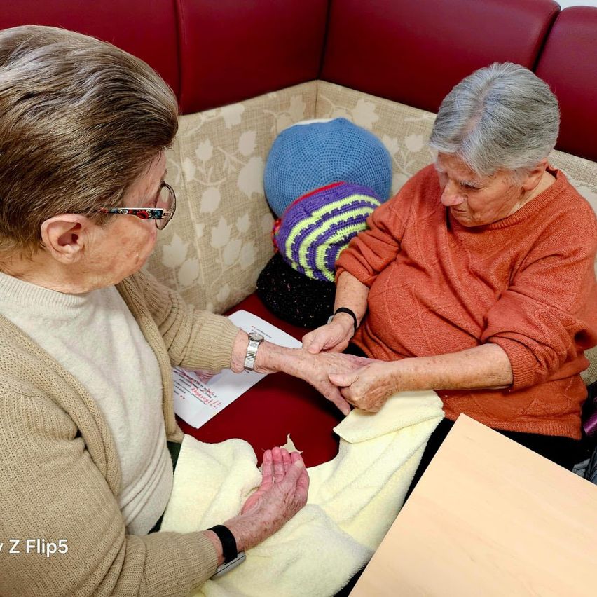 Zwei ältere Frauen sitzen auf einer Couch, eine massiert die Hand der anderen und schaut auf ein Stück Papier.