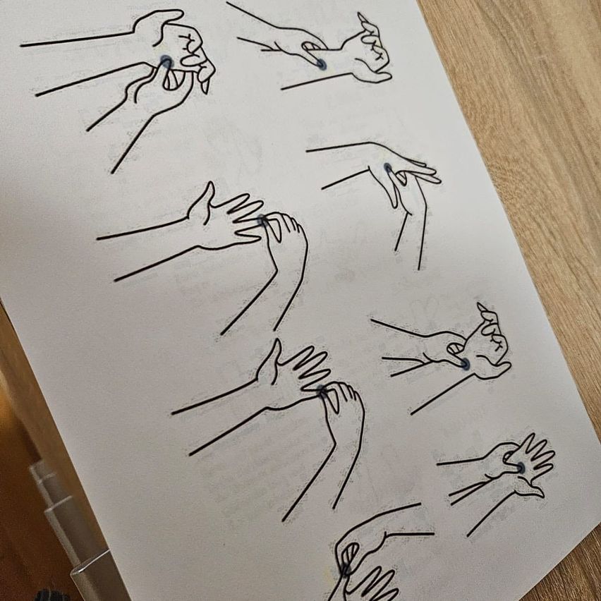 Ein weißes Blatt mit verschiedenen Handhaltungen in Schwarz gezeichnet. Jede Hand hat einen kleinen blauen Punkt am Daumen und Zeigefinger. Die Hände sind auf einer hölzernen Oberfläche positioniert.