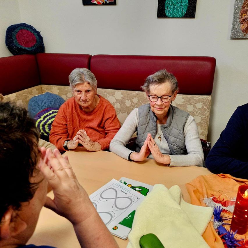 Zwei ältere Frauen sitzen zusammen in einem gemütlichen Raum, tragen Brillen und Pullover. Sie lächeln und klatschen in die Hände. Ein Junge sitzt vor ihnen und hält ein Blatt mit einer Zeichnung. Auf dem Tisch steht eine Kerze.