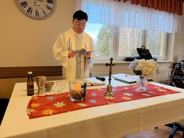 Ein Priester liest aus einem Buch an einem Tisch, der mit einer Blumendekoration und Kerzen bedeckt ist, vor einem Fenster mit weißen Vorhängen.